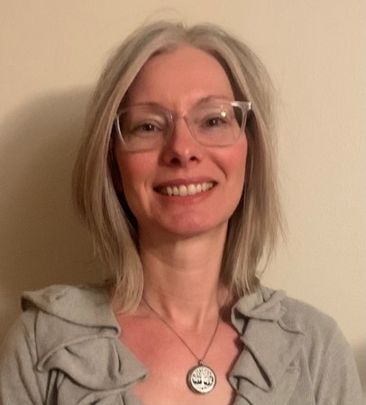 Headshot of Angie Zalig. She is smiling at the camera and is wearing a ruffled grey blouse with a necklace.