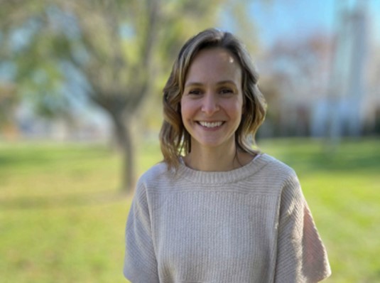 Headshot of Jennifer Shimon. She is wearing a beige sweater and is standing outside in a sunny area with trees, but the background is blurred.