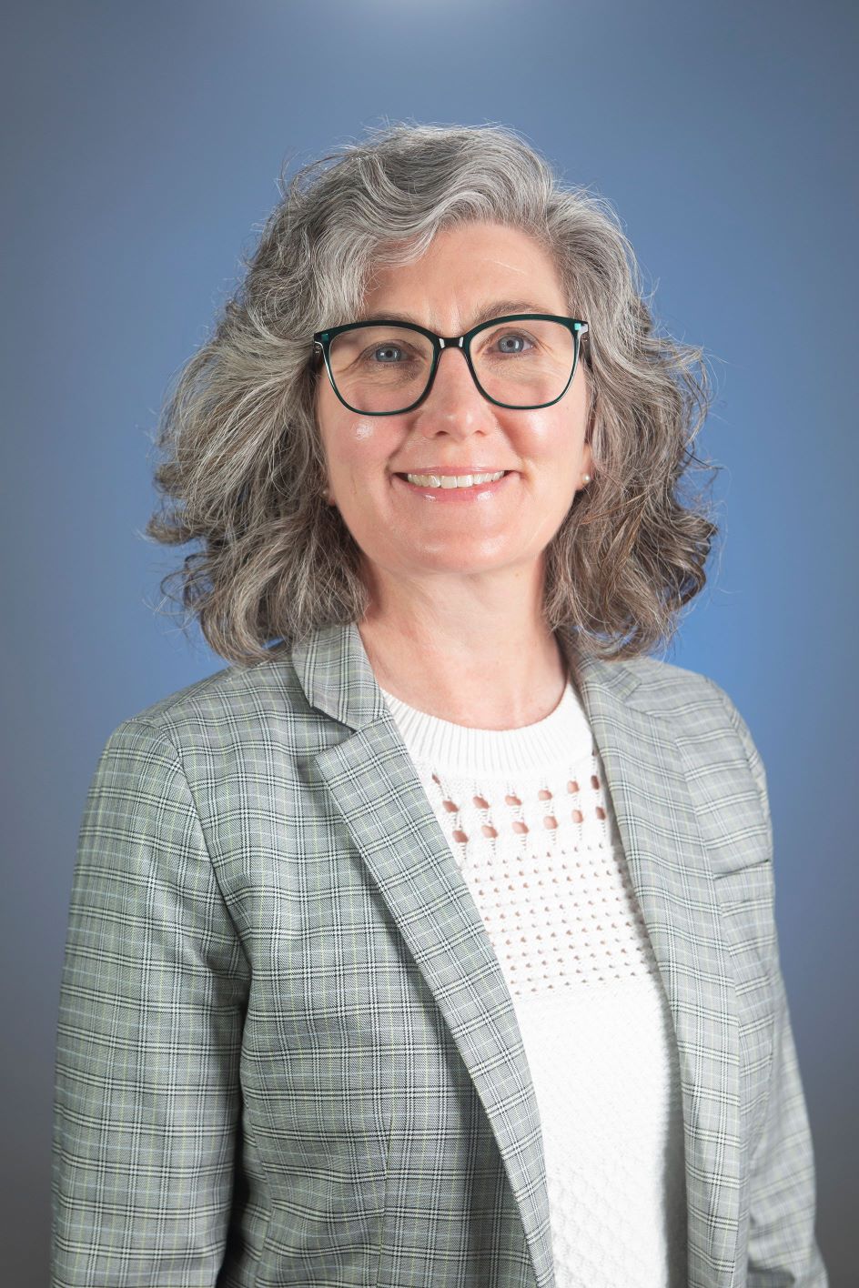 Professional headshot of Kirsten Dorsey. She is smiling in front of a blue background wearing a grey blazer with a white shirt. She wears wide-rimmed glasses, and has blue eyes and grey hair.