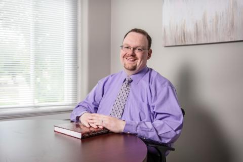 Headshot of Michael Nadorff. He is sitting at a mahogany desk with a psychology book underneath his folded hands.