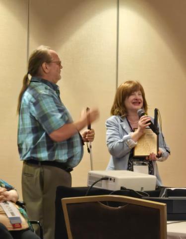 John McMahon and Jennifer Ottowitz stand together in a conference room. Jennifer, holding a microphone and a plaque, smiles while speaking. John beside her holds a white cane and gestures with his hands.