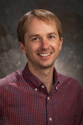 Headshot of Ryan. Ryan has blond hair and is smiling in a red patterned collar shirt.