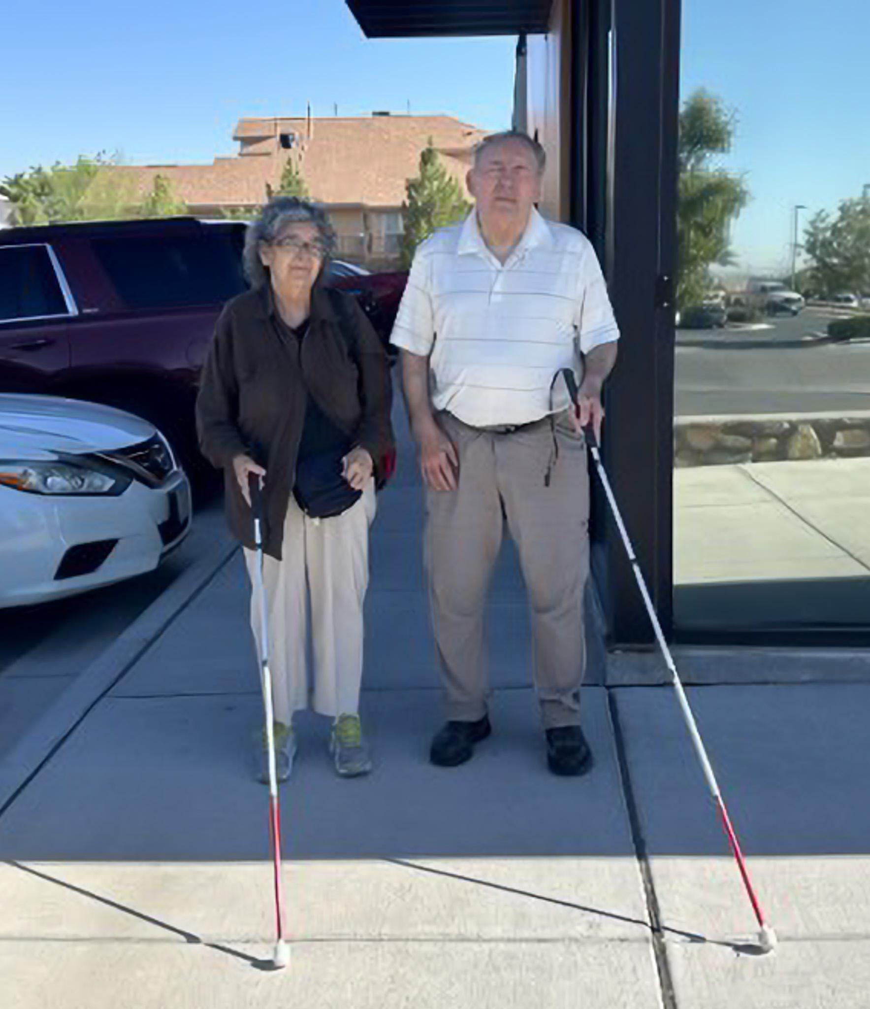 Picture of Jesus. B and his wife standing side by side on a sidewalk. Each one is holding a white cane and looking at the camera.