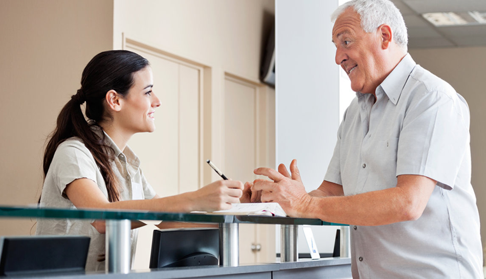 Receptionist holding a pencil and assisting a patient at a desk with papers.