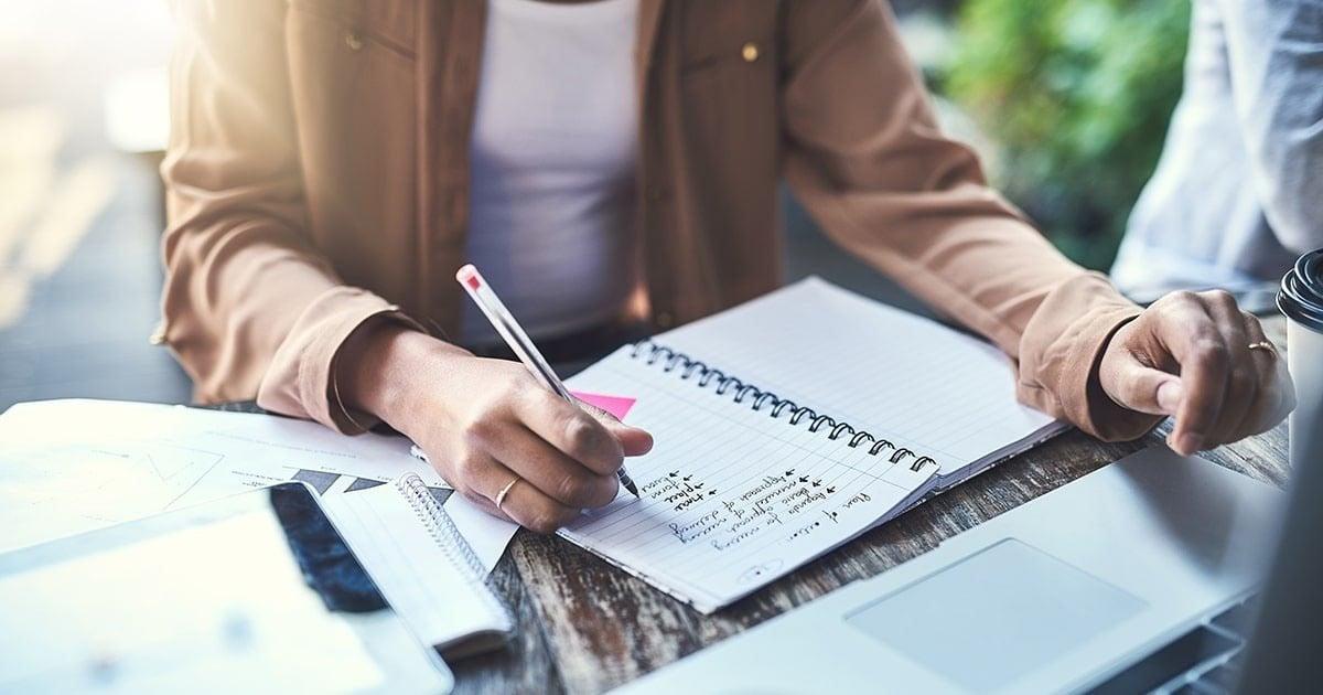 A person writing in a spiral notebook on a wooden table