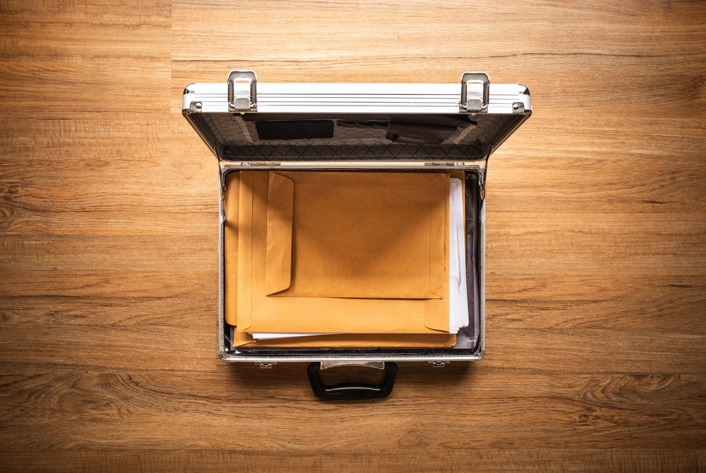 An open silver briefcase with manila folders and papers inside. The briefcase is sitting on top of a wooden table.