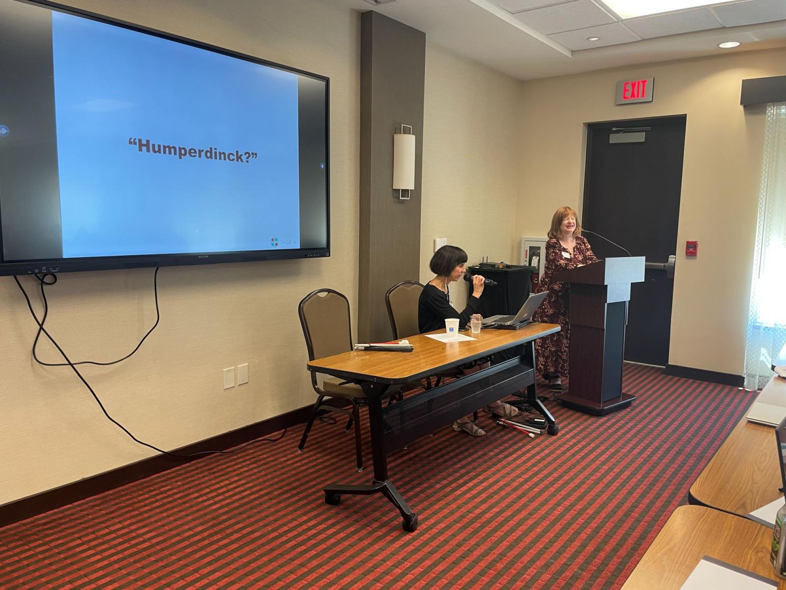 Kendra Farrow and Jennifer Ottowitz standing at the front of a conference room presenting.