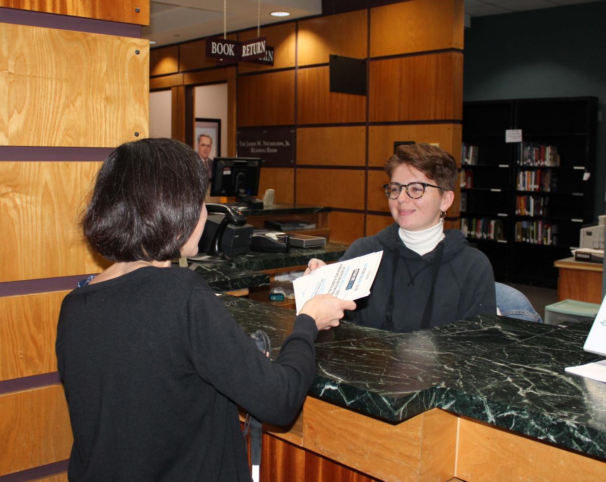 Woman standing passes Time to be Bold material to another person sitting behind a counter in front of her.