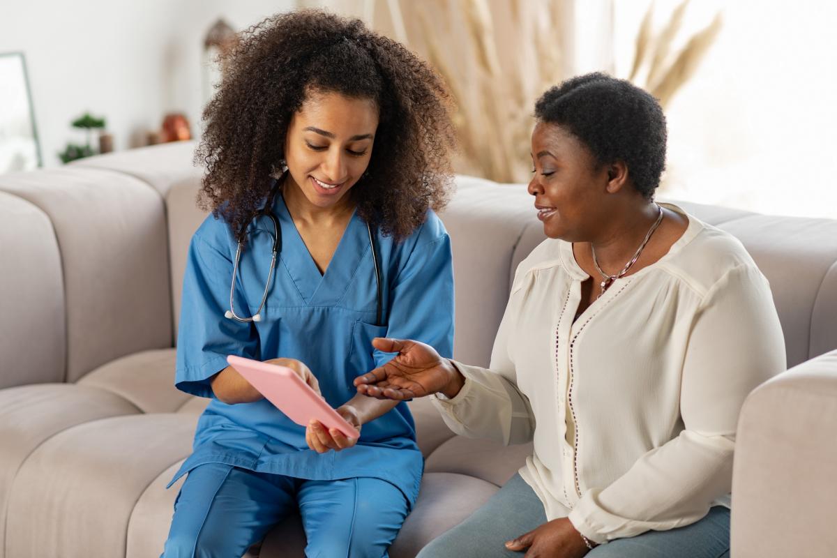 Elderly Black woman talking to a young Black nurse on a grey couch.