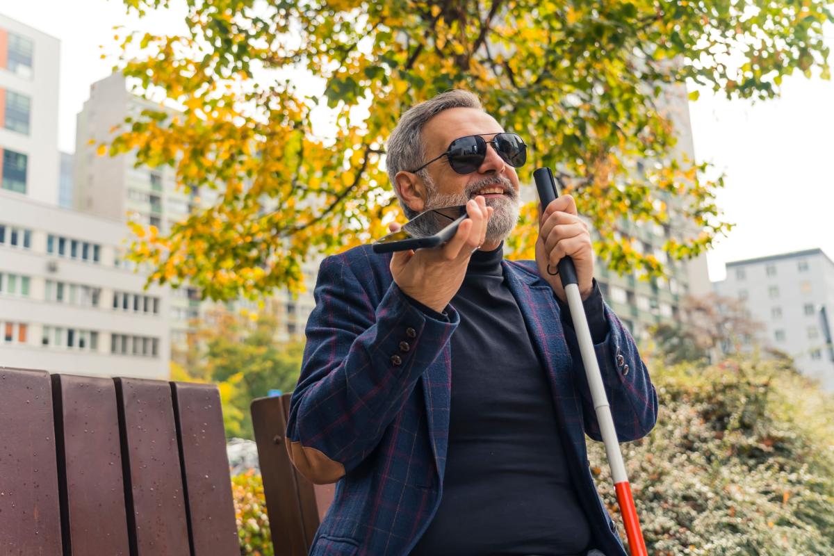 Older man who is blind sits on a park bench holding a white cane while speaking into his phone.