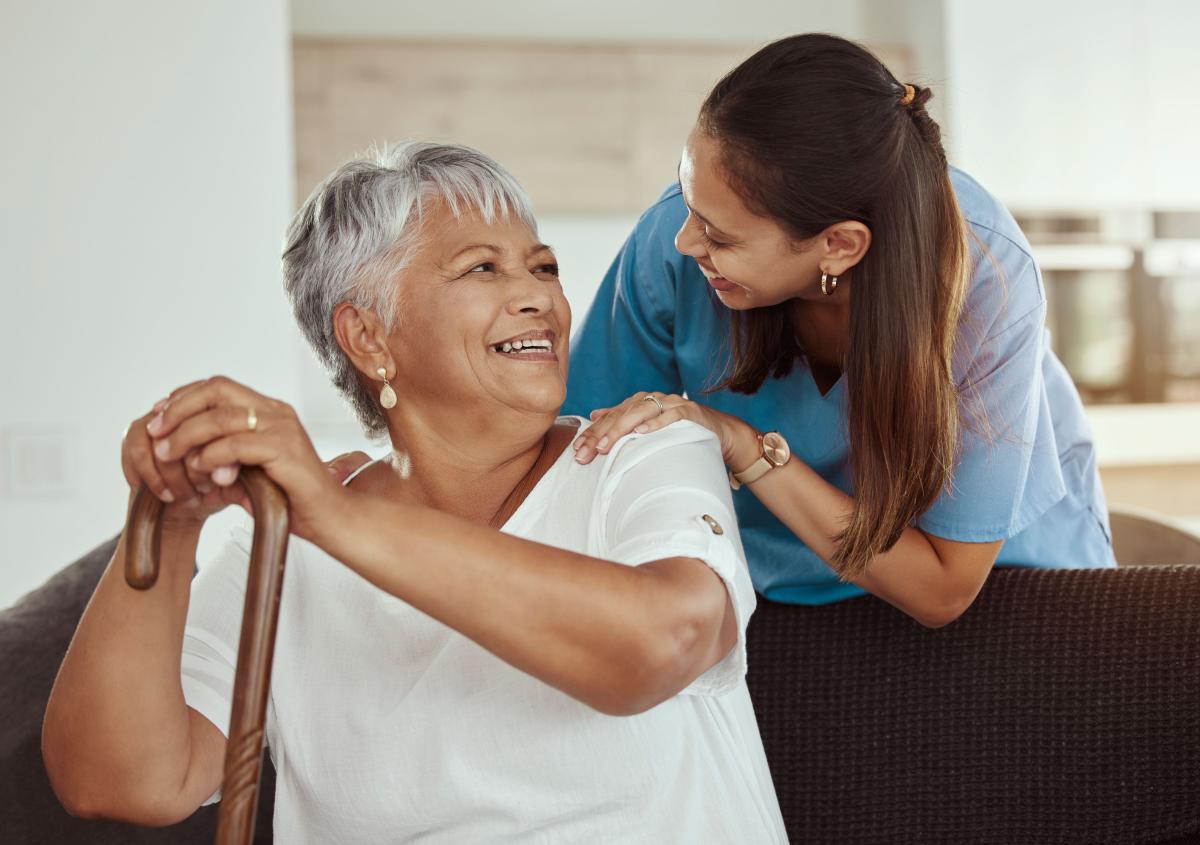 An older woman with a cane is sitting on a couch with her caregiver beside her.