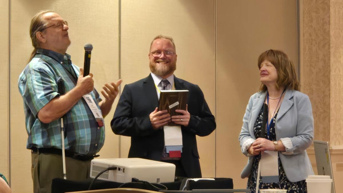 Jennifer Ottowitz, John McMahon, and another person stand at the front of a conference room. John on the left holds a microphone and gestures as he speaks, while the man in the center, dressed in a suit, smiles and holds a plaque. Jennifer is on the right, holding a white cane, and looks toward the speaker.